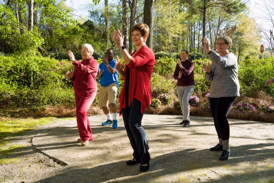 A group of five seniors practicing Tai Chi outdoors on a sunny day, surrounded by green trees and bushes with some flowers in the background.