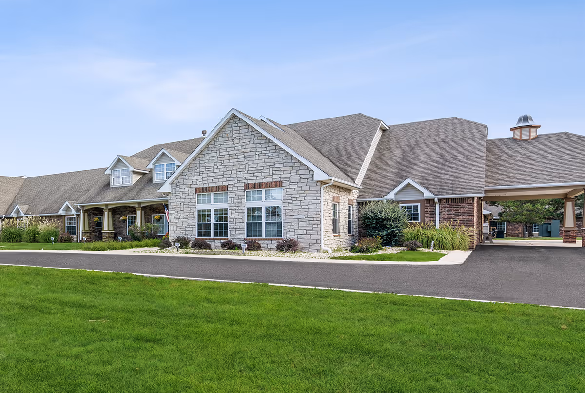 Exterior view of a single-story senior living facility building with stone and brick facade, multiple windows, a covered entrance, and a well-maintained lawn and landscaping under a clear blue sky.