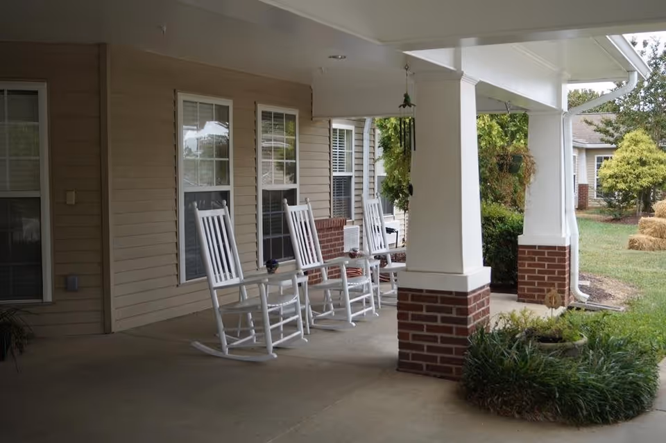 Covered porch area with three white rocking chairs and small tables in front of a beige building with windows. There are plants and greenery around the porch and a grassy yard with some bushes and hay bales in the background.