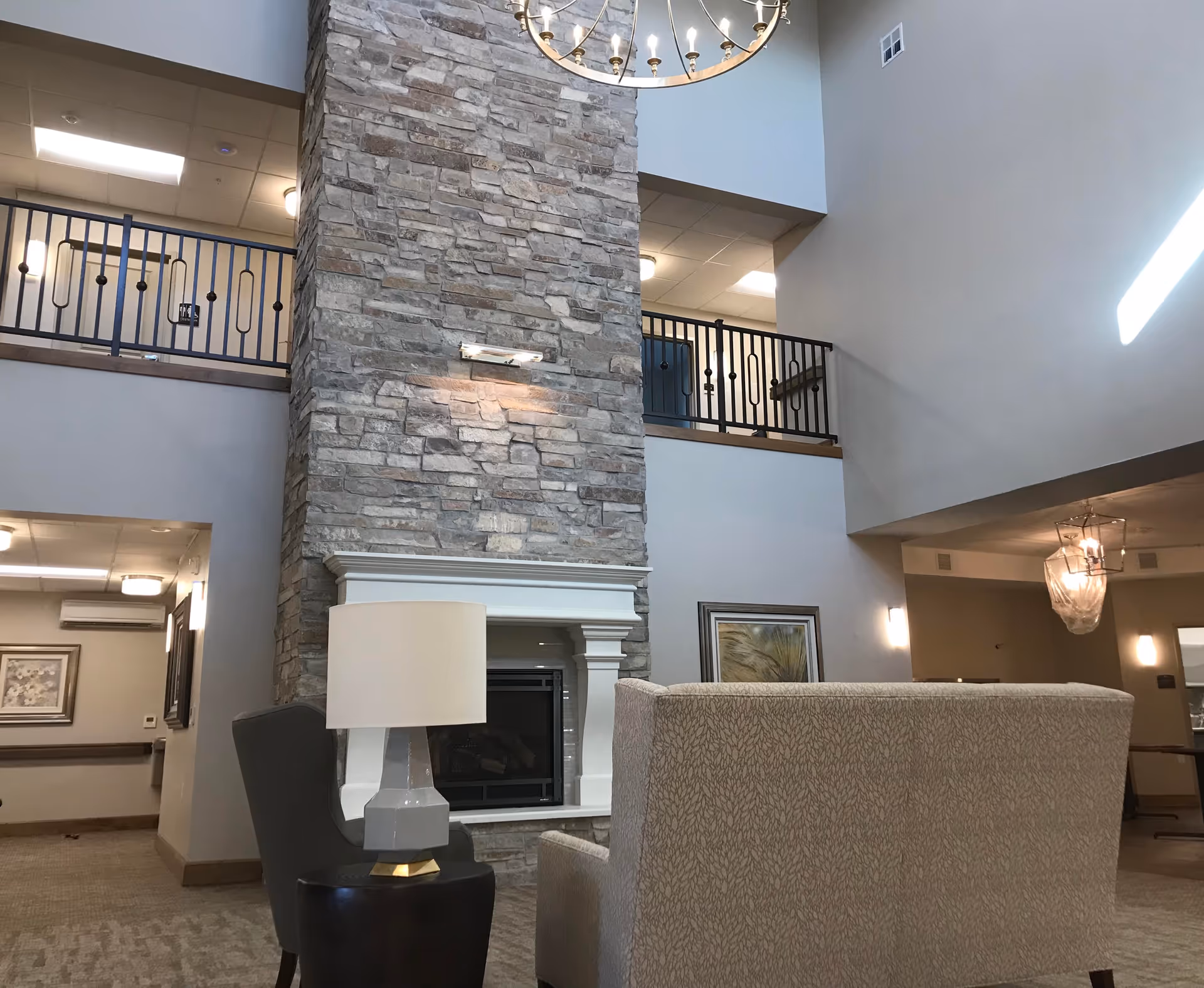 Interior view of a senior living facility lounge area featuring a tall stone fireplace with a white mantel. In front of the fireplace are a beige patterned sofa and a gray armchair with a modern table lamp on a small round table. The space has high ceilings with a chandelier, a second-floor balcony with black railings, and neutral-colored walls with framed artwork.