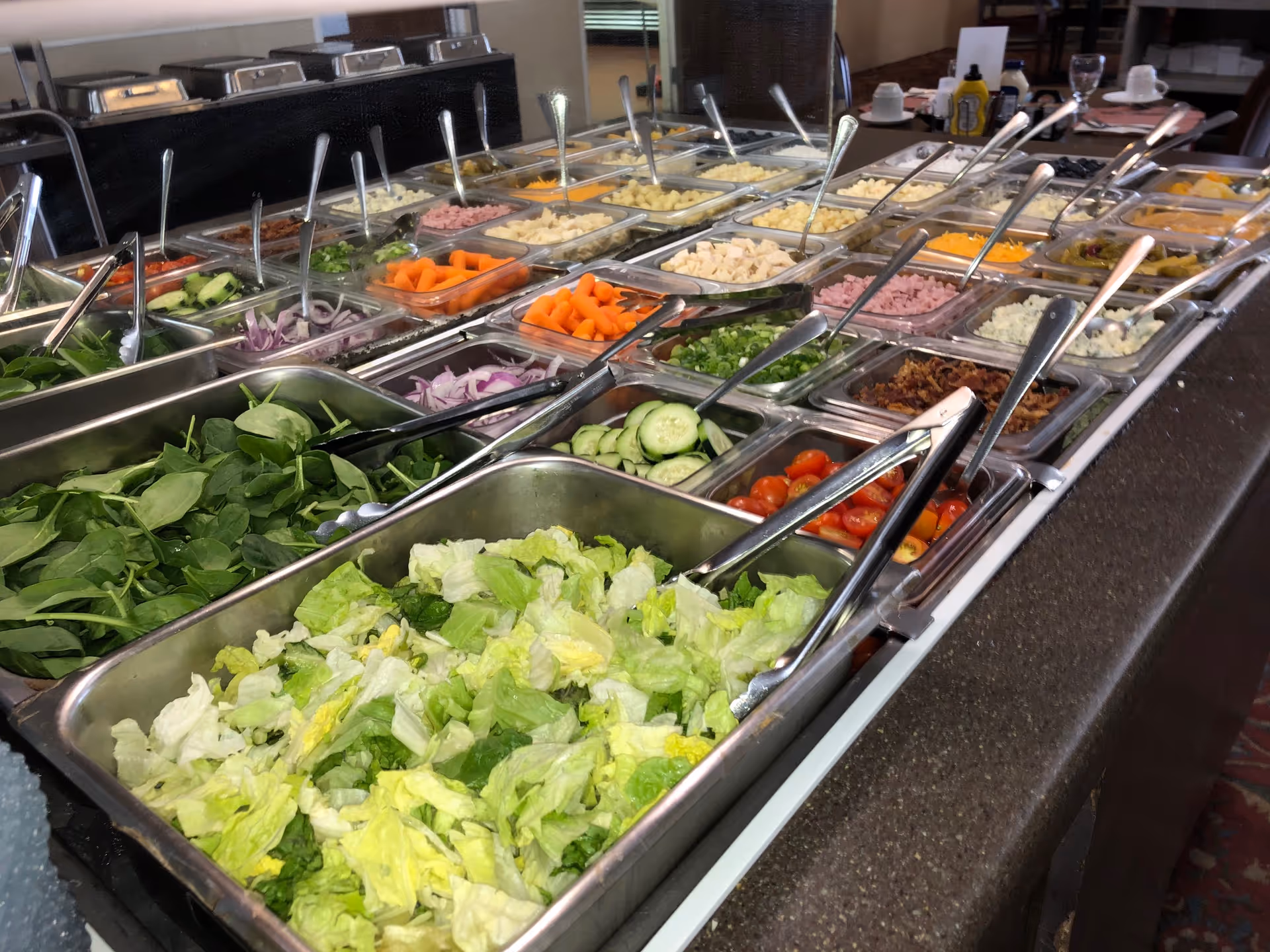 A salad bar with various fresh vegetables and toppings including lettuce, spinach, cucumbers, cherry tomatoes, carrots, onions, bacon bits, cheese, and dressings, all arranged in metal and plastic containers with serving utensils.