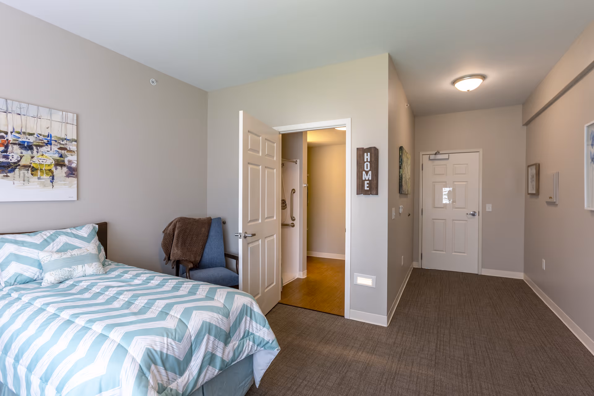 A bedroom with a bed covered in a light blue and white chevron patterned comforter, a blue chair with a brown blanket draped over it, a painting of boats on the wall, an open door leading to a bathroom, and a hallway with a closed white door at the end. A wooden sign with the word 'HOME' is mounted on the wall near the bathroom door.