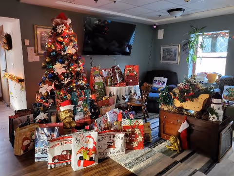 A festive living room decorated for Christmas with a decorated Christmas tree surrounded by numerous gift bags and presents. The room features a wall-mounted TV, a window with a patterned curtain, a wooden chest adorned with holiday decorations including a stocking and a snowman figure, and comfortable seating. The space is warmly lit and has a cozy holiday atmosphere.