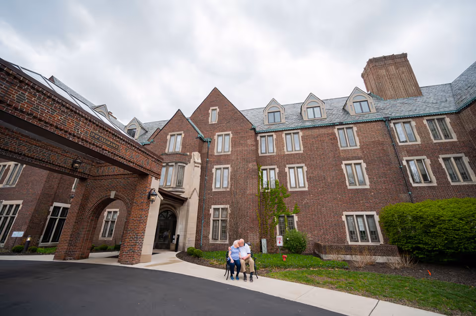 An elderly couple sitting on a bench outside a large brick building with multiple windows and a slate roof, under a cloudy sky. The building has a covered entrance with an archway and a sign indicating 13' 9" clearance.