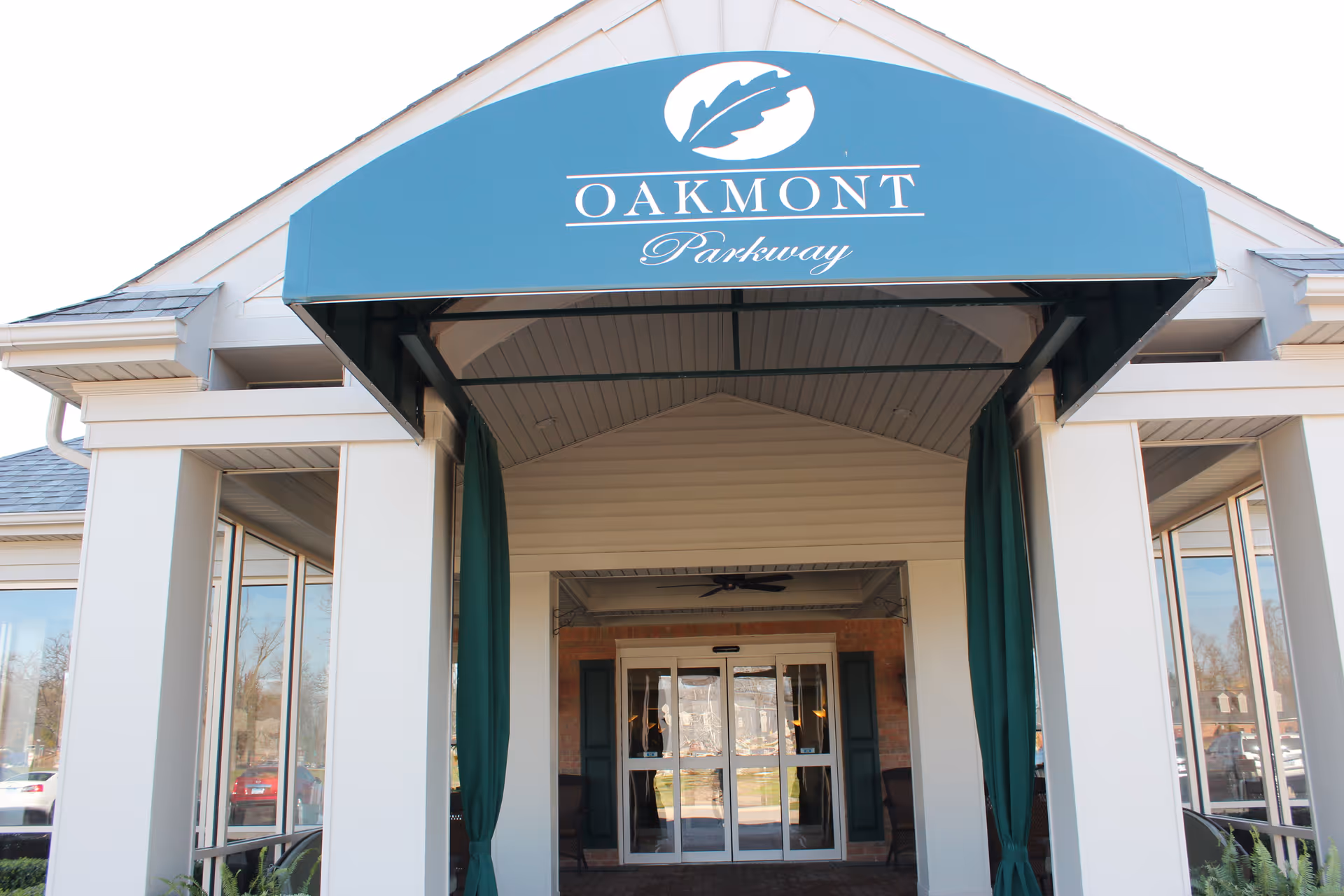 Entrance of Oakmont Parkway Senior Community featuring a covered walkway with a blue awning displaying the community's name and logo above glass double doors.