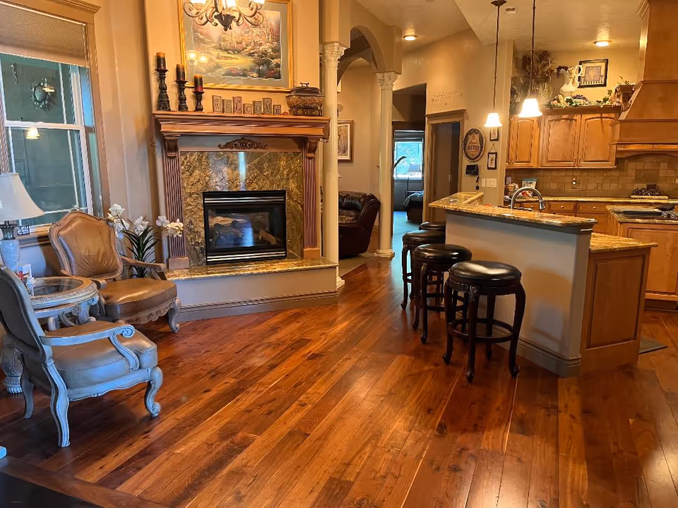 Interior view of a cozy living space featuring a wooden floor, a fireplace with a decorative mantle, two upholstered chairs with a small round table, and a kitchen area with a granite countertop and four bar stools. The kitchen has wooden cabinets and pendant lights hanging above the counter. There is a hallway leading to other rooms in the background.