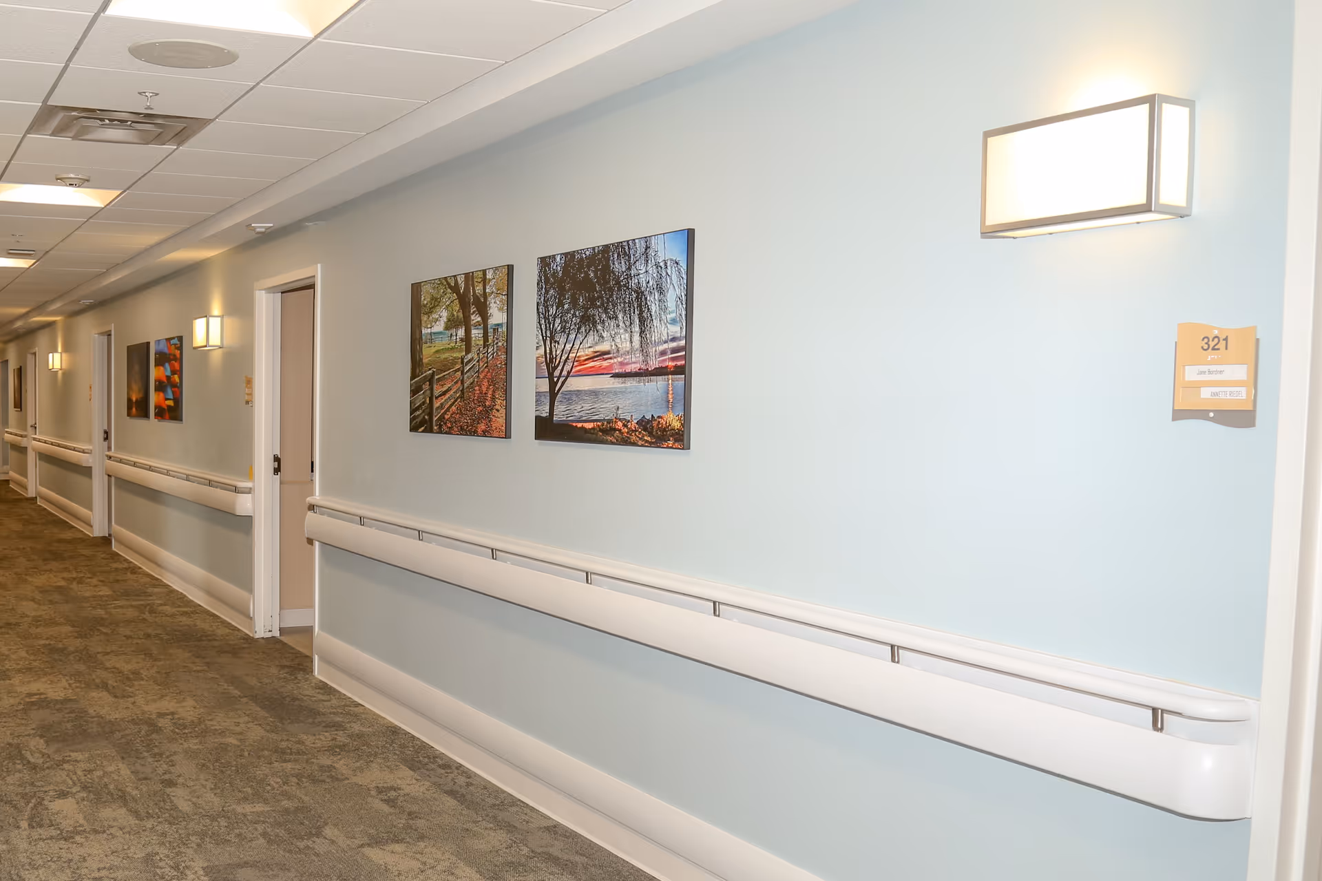 Well-lit hallway of a senior living facility with handrails, room doors, and framed landscape artwork on pale blue walls.