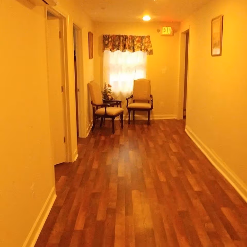 A hallway with wooden flooring and cream-colored walls, featuring two upholstered chairs placed near a window with floral curtains. There are doorways on both sides of the hallway and framed pictures on the walls. An illuminated exit sign is visible above the window.