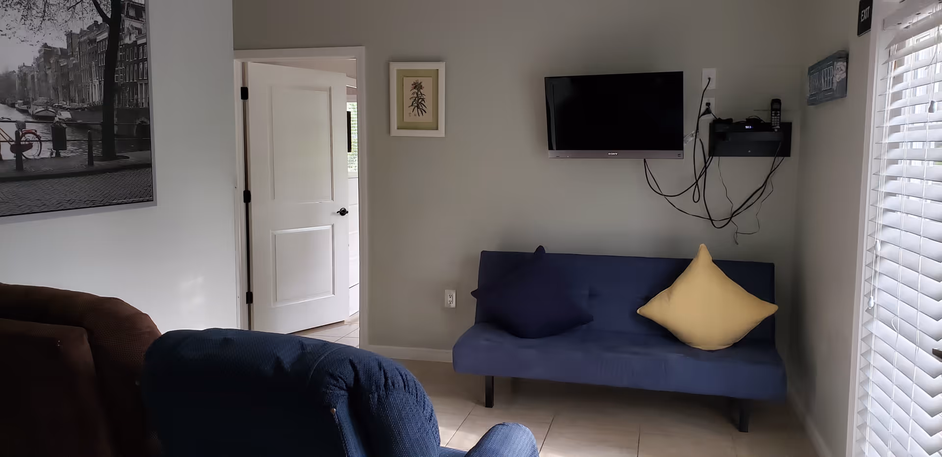 A small living room area with a blue futon sofa featuring two cushions, one dark blue and one yellow. A flat-screen TV is mounted on the wall above the sofa with visible cables hanging down. To the left, there is an open white door leading to another room. A framed picture hangs on the wall next to the door, and a large black and white photo is partially visible on the left wall. The room has tiled flooring and a window with white blinds on the right side.