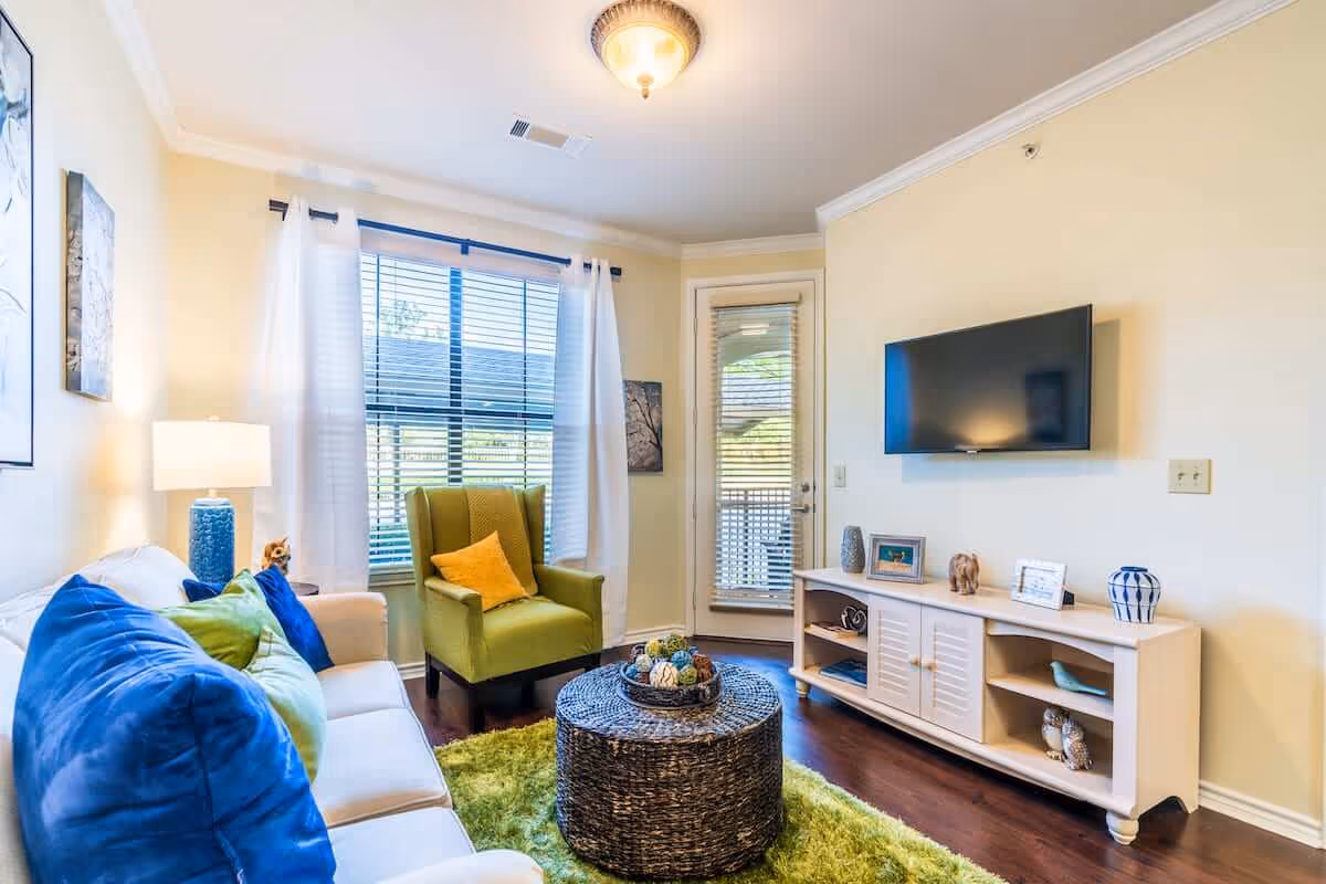Bright living room with a sofa, blue and green pillows, a green armchair, wall-mounted TV, and a wicker coffee table on a green rug.