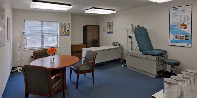 A medical examination room with a round wooden table and four chairs, a medical examination table with a teal cushion, a stool, a skeleton model near the window, medical posters on the walls, and a desk with a chair in the background.