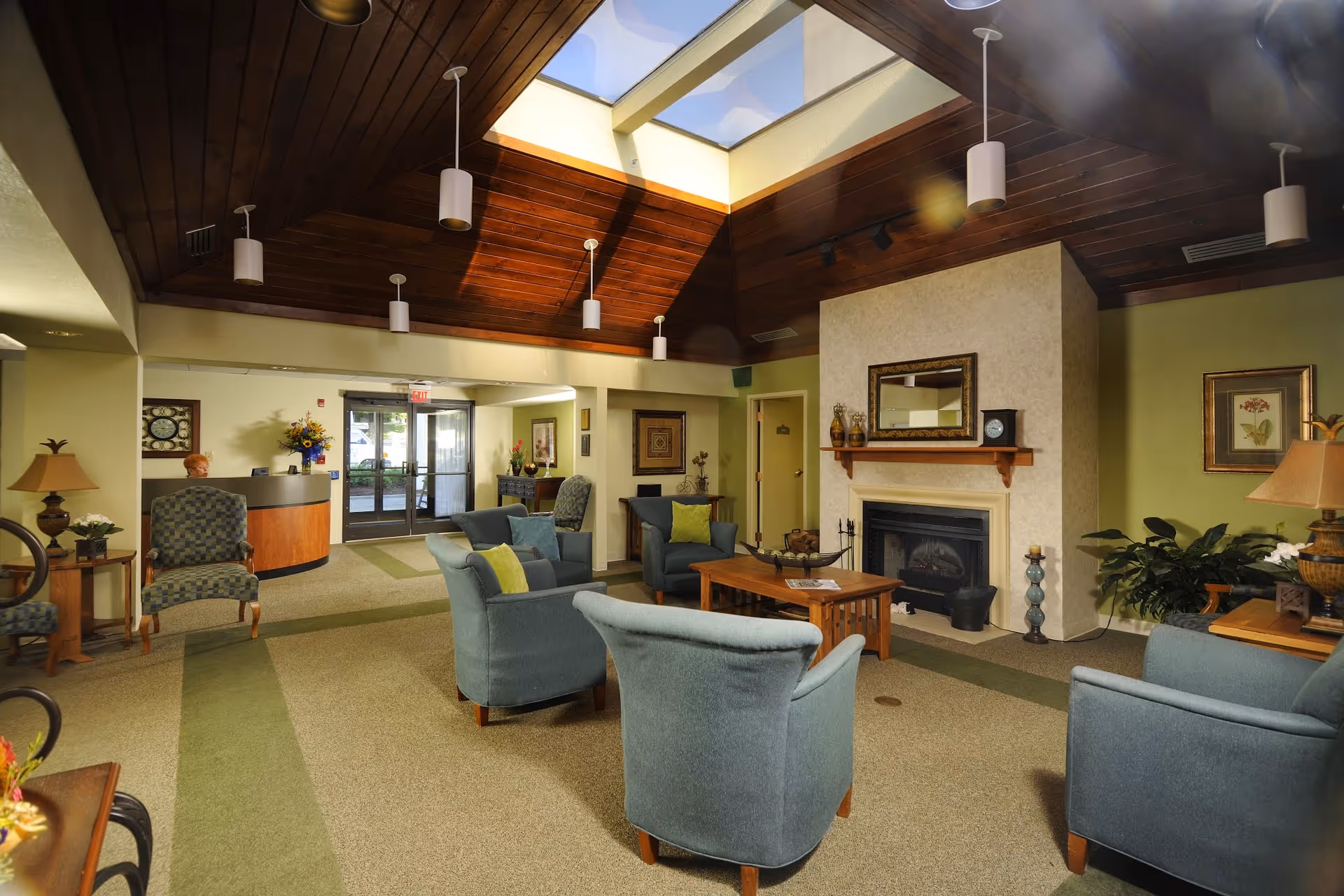 A cozy senior living facility common area with comfortable blue armchairs and a wooden coffee table arranged around a fireplace. The room has a high wooden ceiling with a skylight, soft lighting from hanging fixtures, and decorative plants and artwork on the walls. A reception desk is visible near the entrance with glass doors leading outside.