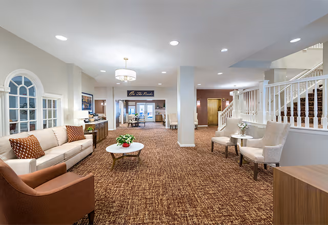 Bright, carpeted senior living common area with sofas, armchairs, a coffee table, and a staircase.