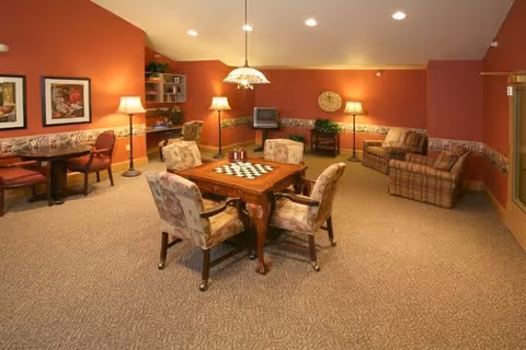 A cozy common area with warm orange walls and beige carpet. The room features a wooden table with a chessboard and four cushioned chairs around it. There are additional seating areas with armchairs and a small table with a lamp. A TV sits on a stand against the far wall, and framed artwork and a clock decorate the walls. Soft lighting is provided by ceiling lights and several lamps.