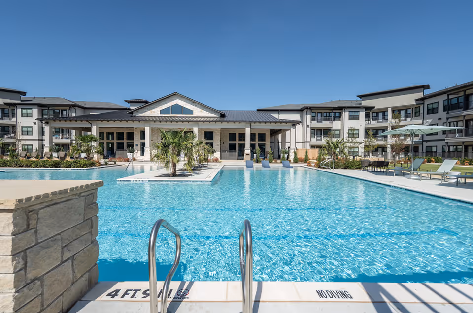Outdoor swimming pool with a ladder, lounge chairs, and umbrella in front of a multi-story senior living building under a clear blue sky.