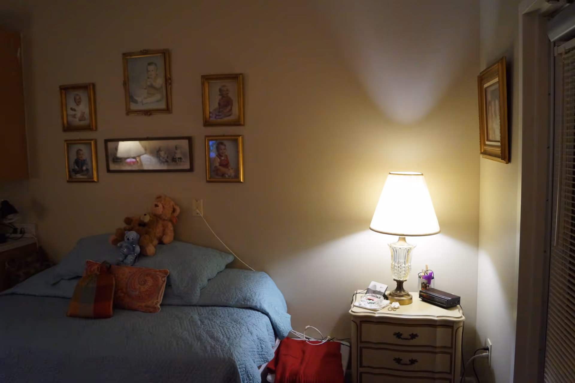 A cozy bedroom with a bed covered in a blue quilt and several pillows, along with stuffed teddy bears placed on the bed. Above the bed, there are six framed pictures of babies arranged on the wall. To the right of the bed is a small nightstand with a lit table lamp, a clock radio, and some small items. The room has a warm and inviting atmosphere.