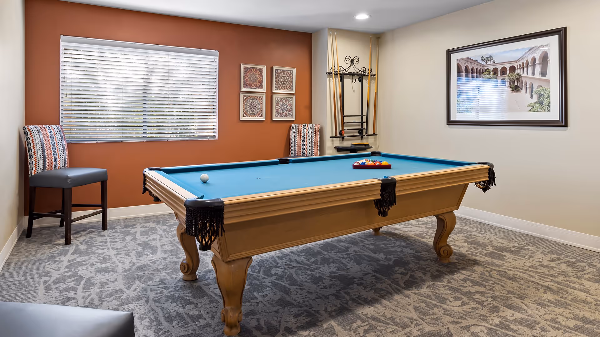 A well-lit room with a pool table in the center, featuring a blue felt surface and a set of billiard balls arranged in a triangle. The room has a window with blinds, two patterned chairs against an orange accent wall, a wall-mounted rack holding pool cues, and a framed picture on a beige wall. The floor is carpeted with a patterned design.