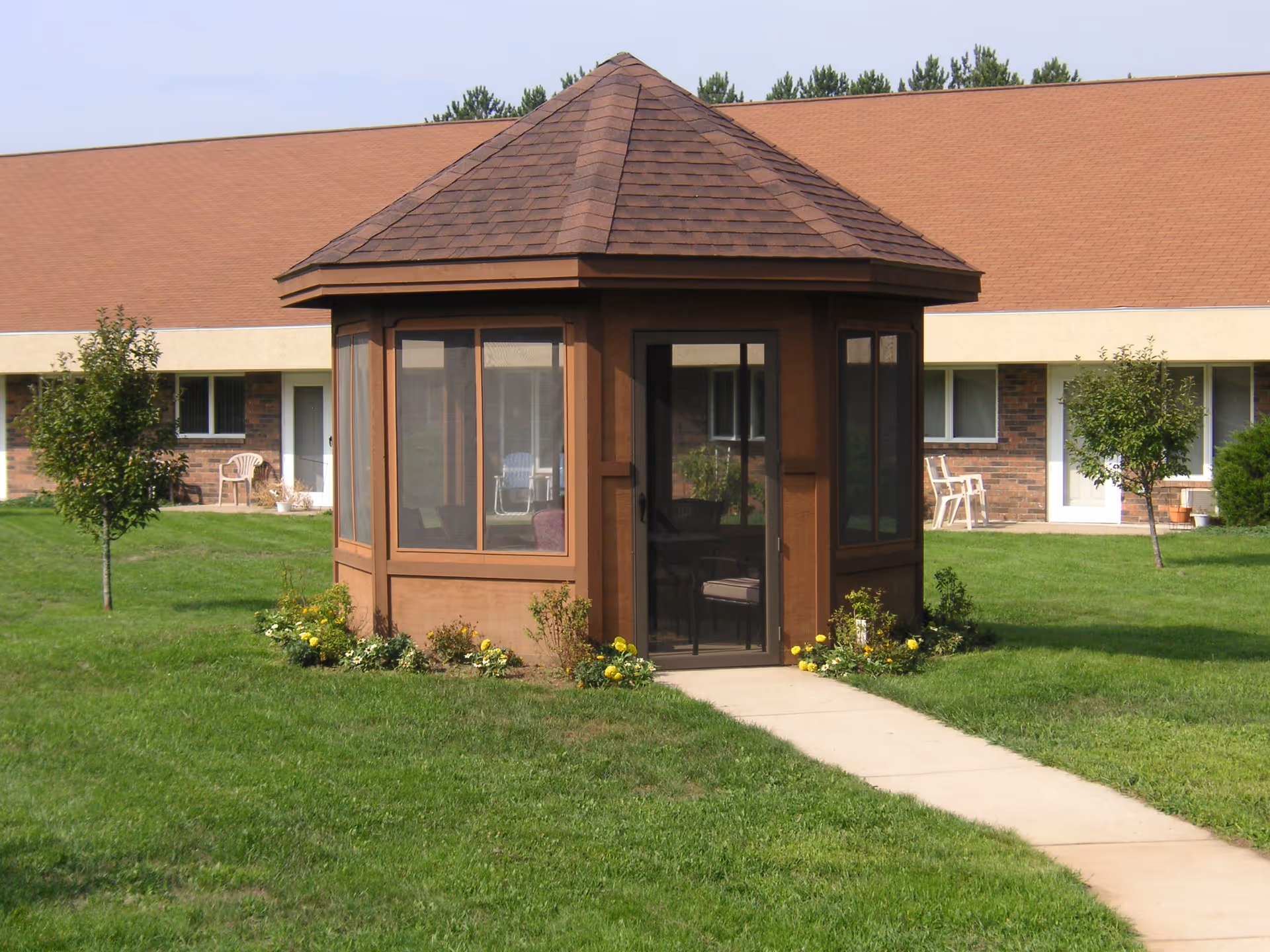 A small brown gazebo with a shingled roof and screened windows stands on a concrete path surrounded by green grass and small flower beds. In the background, there is a single-story brick building with white doors and windows, and a few plastic chairs placed outside.