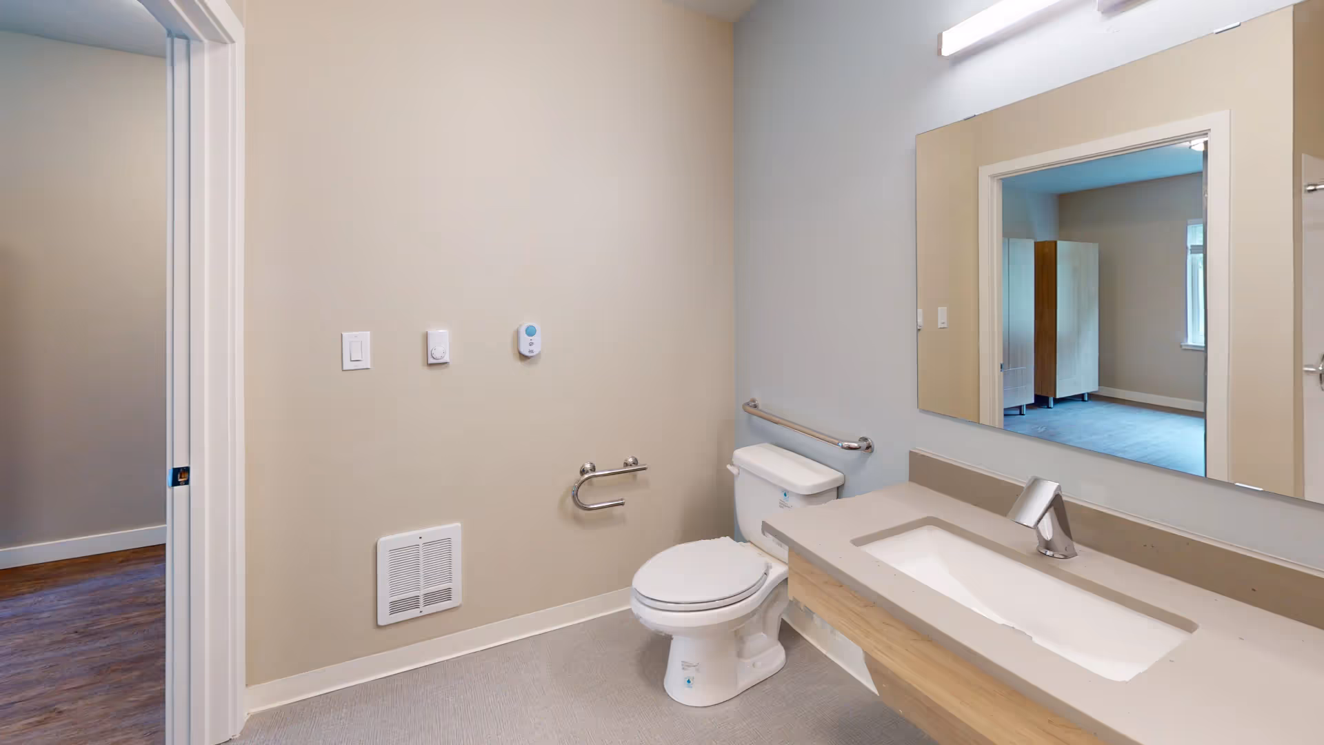 Accessible bathroom with a toilet, wall grab bars, a sink vanity and a large mirror reflecting an adjacent room.
