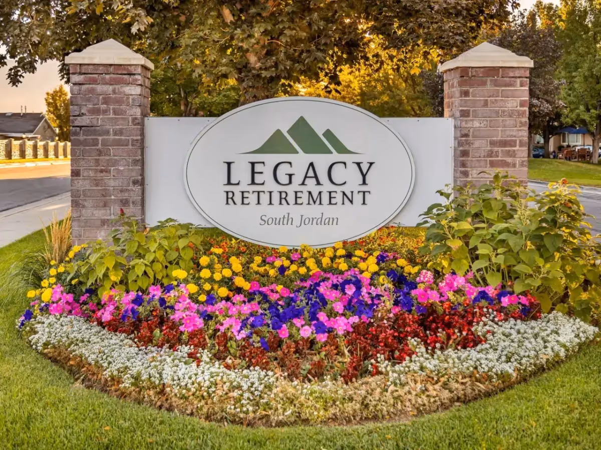 A landscaped entrance sign for Legacy Retirement South Jordan, featuring a white oval sign with green mountain graphics and black text, flanked by two brick pillars and surrounded by colorful flowers and green grass.