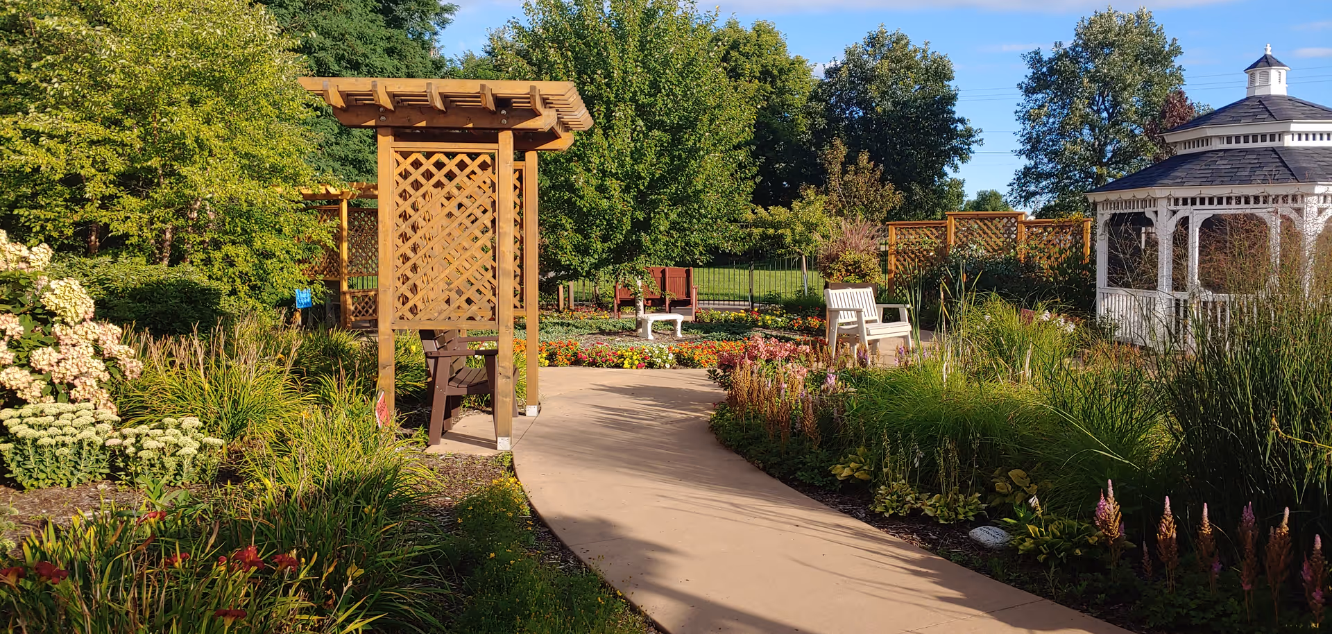 A sunny outdoor garden area with a paved walkway winding through lush greenery, colorful flowers, and wooden trellises. There are several benches along the path and a white gazebo on the right side. Trees and a clear blue sky are visible in the background.
