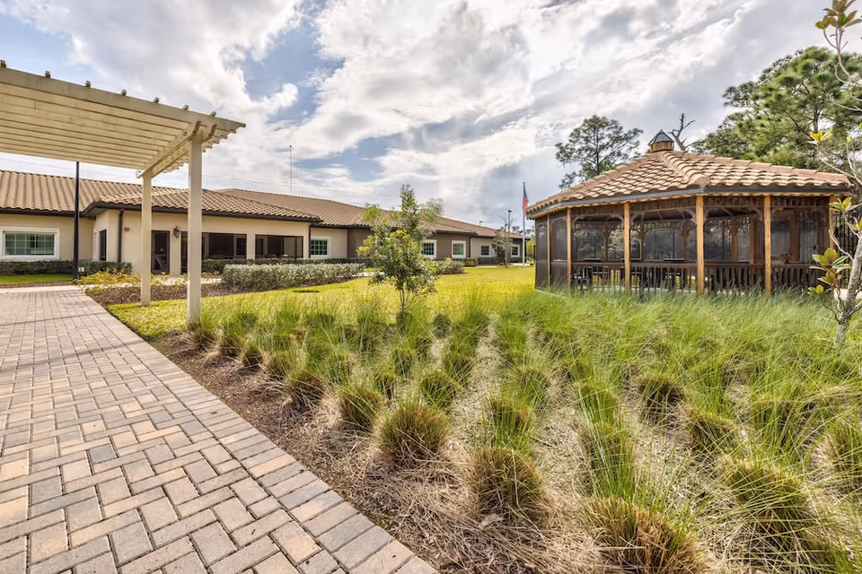 Landscaped courtyard with a gazebo, paved walkway, and a single-story building under a partly cloudy sky.