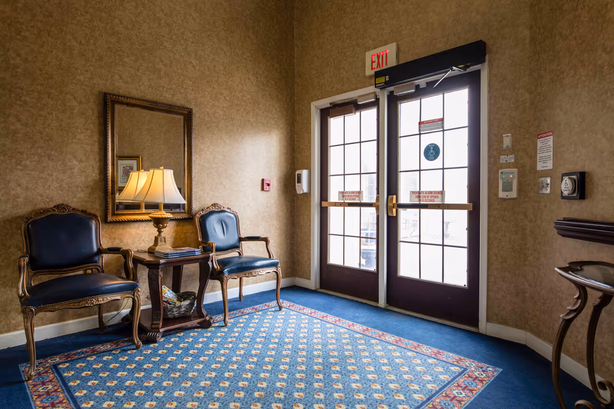 A small seating area near a set of double glass exit doors. The area features two ornate wooden chairs with dark blue cushions, a wooden side table with a lamp and some magazines, a large mirror on the wall, and a patterned blue carpet. The walls are covered in a textured beige wallpaper.