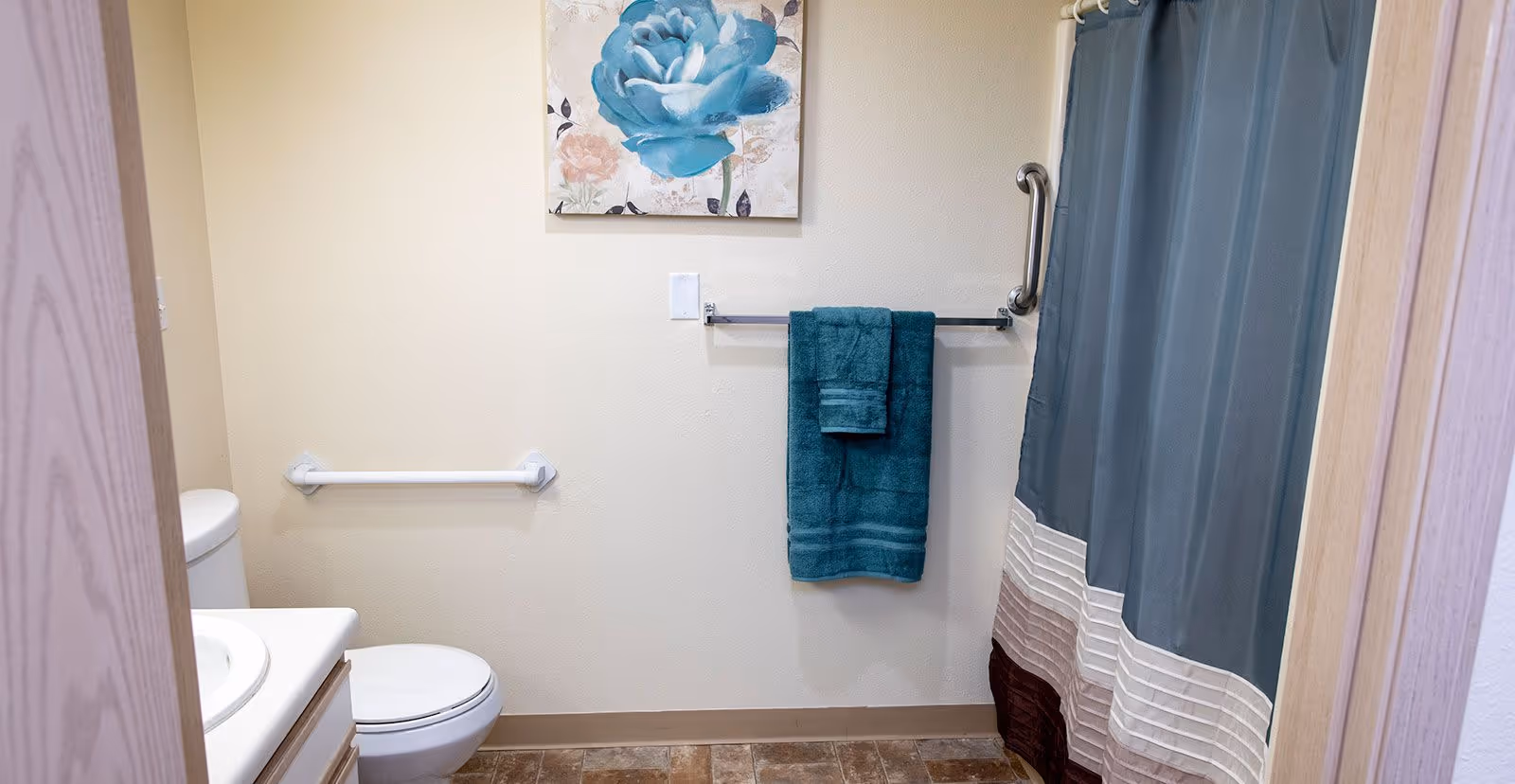 A small bathroom with a toilet, a sink countertop, a towel rack holding two teal towels, a shower curtain in shades of blue and brown, a grab bar next to the shower, and a wall-mounted painting of a blue flower.