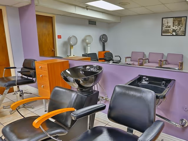 Interior salon area with black styling chairs, shampoo sinks, hooded hair dryers and a mirrored wall with seating.