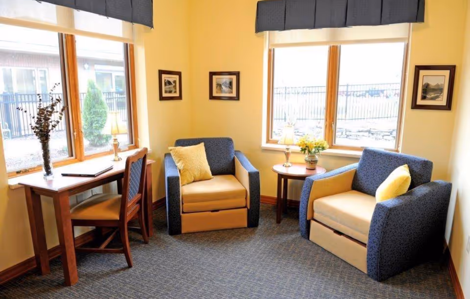 A cozy sitting area with two armchairs featuring blue and beige upholstery, each with a yellow pillow. Between the chairs is a small round wooden table with a vase of yellow flowers and a lamp. To the left, there is a wooden desk with a chair, a lamp, and a vase with dried branches. The room has two large windows with dark valances, allowing natural light to brighten the space. The walls are painted light yellow and decorated with framed pictures.