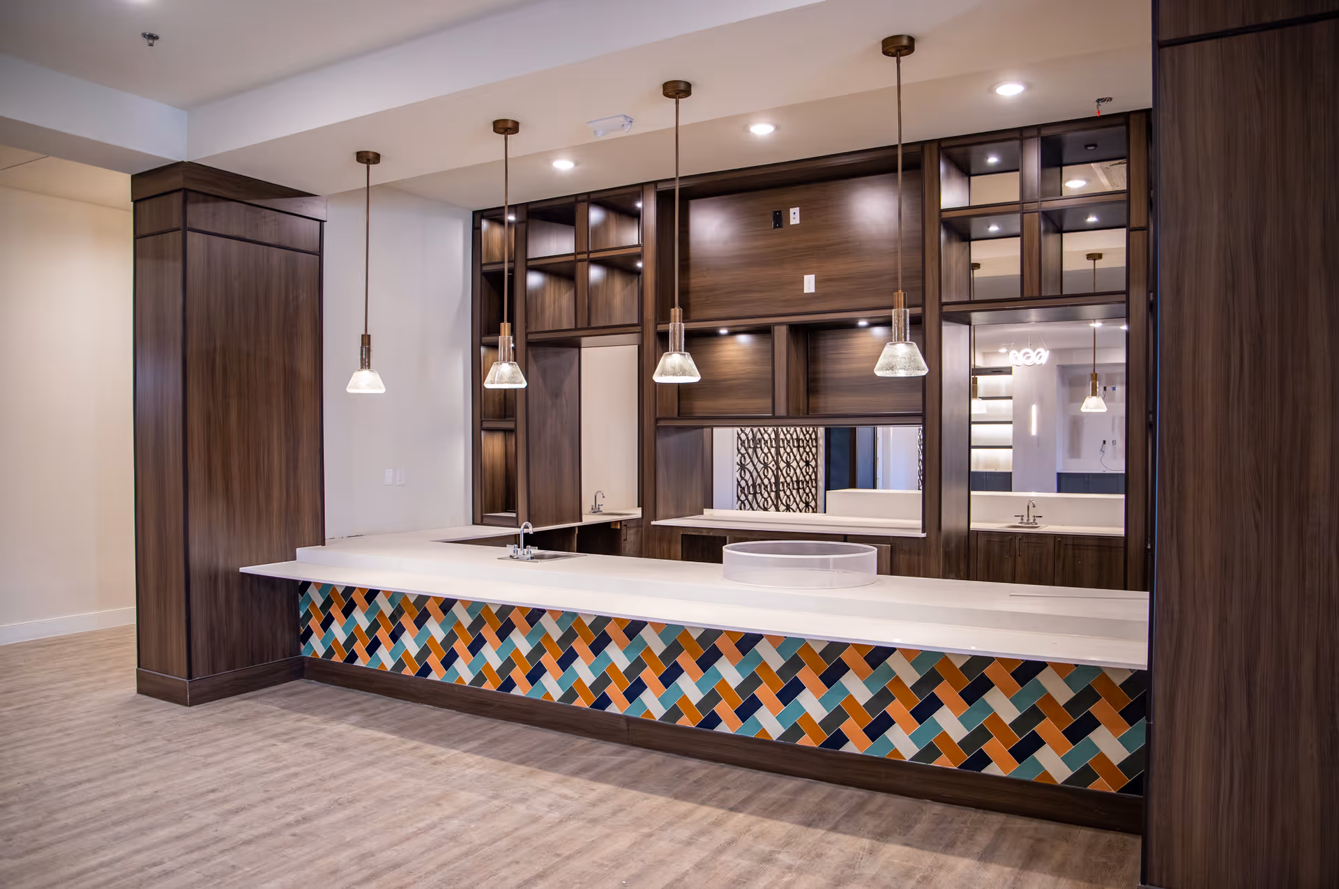 Modern interior space featuring a long white countertop with a colorful geometric patterned front panel. Above the counter, five pendant lights hang from the ceiling. The back wall has dark wood cabinetry with open shelves and mirrors, reflecting parts of the room. The floor is light wood, and the overall design is clean and contemporary.