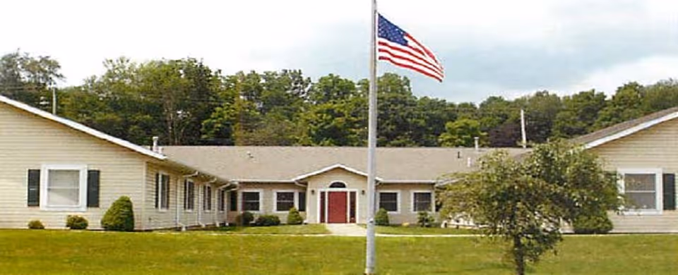 Single-story beige building with a central entrance and red door, surrounded by green grass and trees in the background. An American flag on a flagpole is prominently displayed in front of the building.
