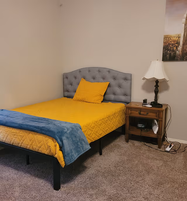 Simple bedroom with a gray tufted headboard, yellow bedspread and pillow, a wooden nightstand with a lamp, and carpeted floor.