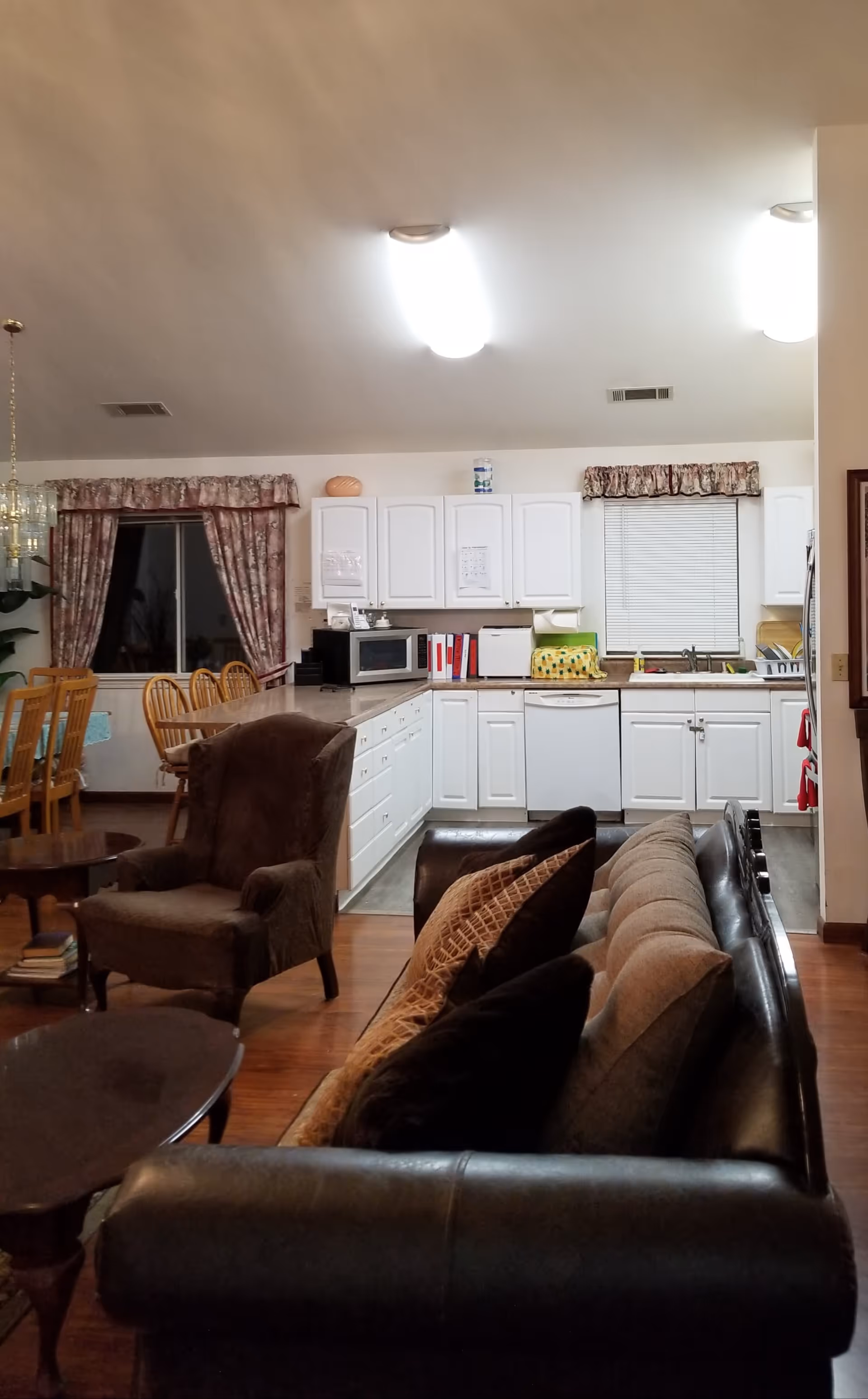 Interior view of a senior living facility room showing a living area with a dark leather sofa and a brown armchair, a wooden coffee table, and a dining area with wooden chairs and a table covered with a blue tablecloth. In the background, there is a kitchen with white cabinets, a microwave, a dishwasher, a sink, and windows with floral curtains and blinds.