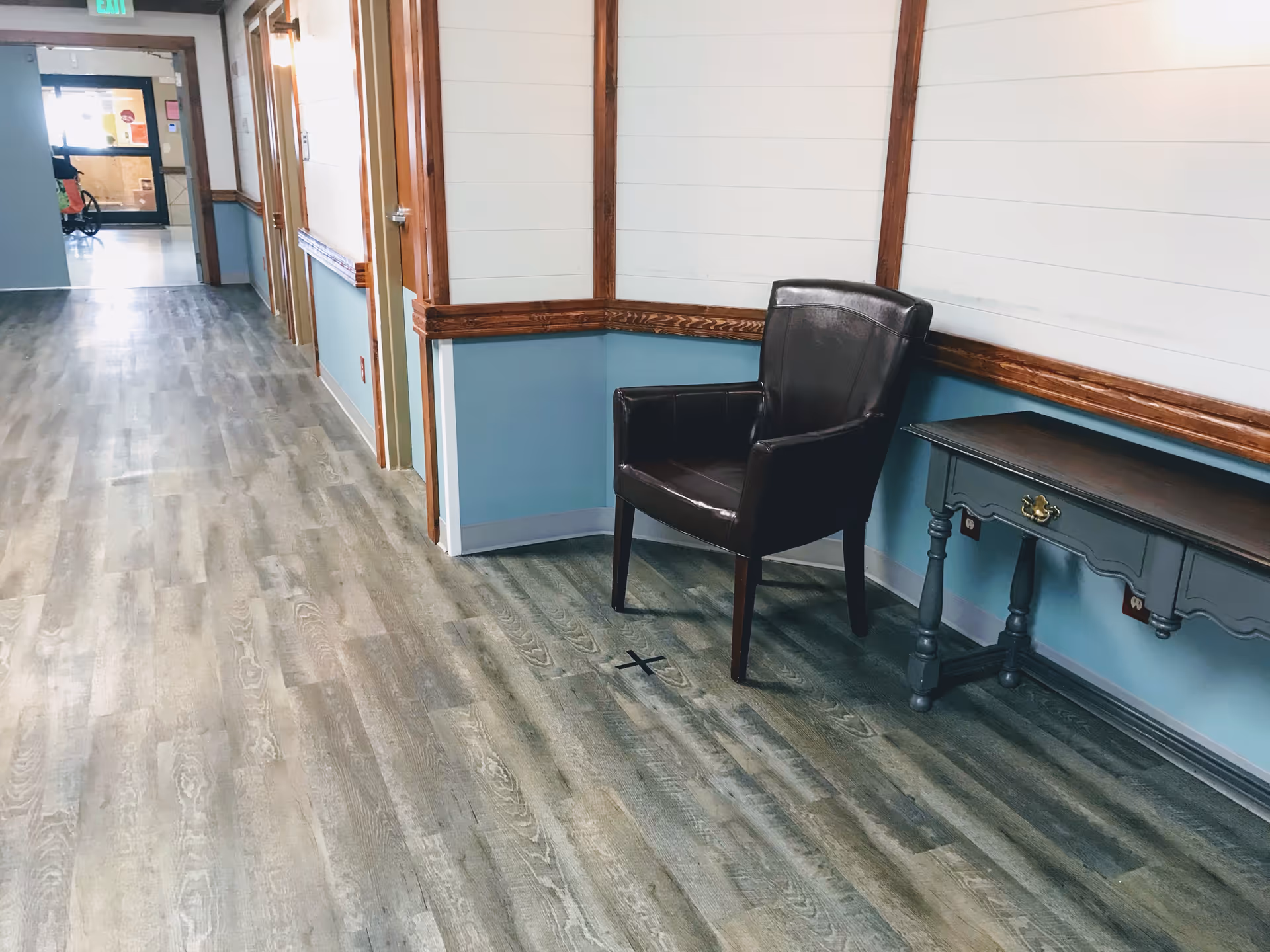 A clean hallway with wood-patterned flooring and light blue walls with wooden trim. There is a black leather chair and a small blue wooden table with a drawer along the right wall. The hallway leads to a door with a glass window at the far end.