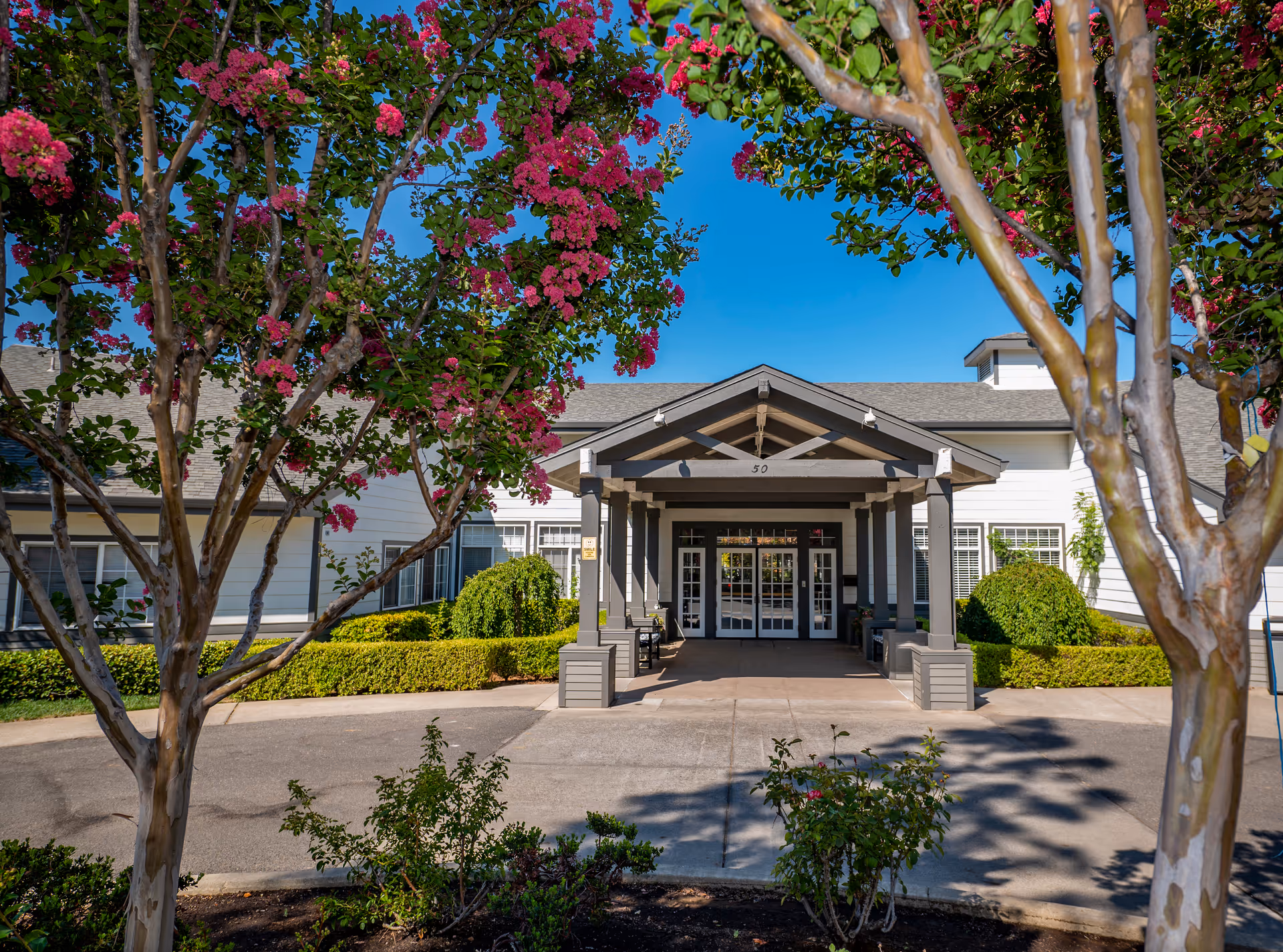 Front exterior view of a single-story building with a covered entrance supported by columns. The building is white with gray trim and surrounded by neatly trimmed bushes and flowering trees with pink blossoms. The sky is clear and blue.