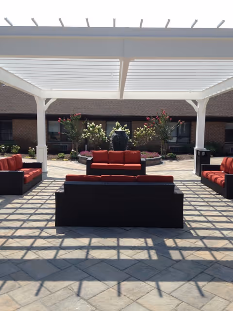 Outdoor seating area with three dark wicker sofas featuring red cushions arranged under a white pergola. In the background, there is a garden with flowering plants and a water fountain, with a brick building behind it.
