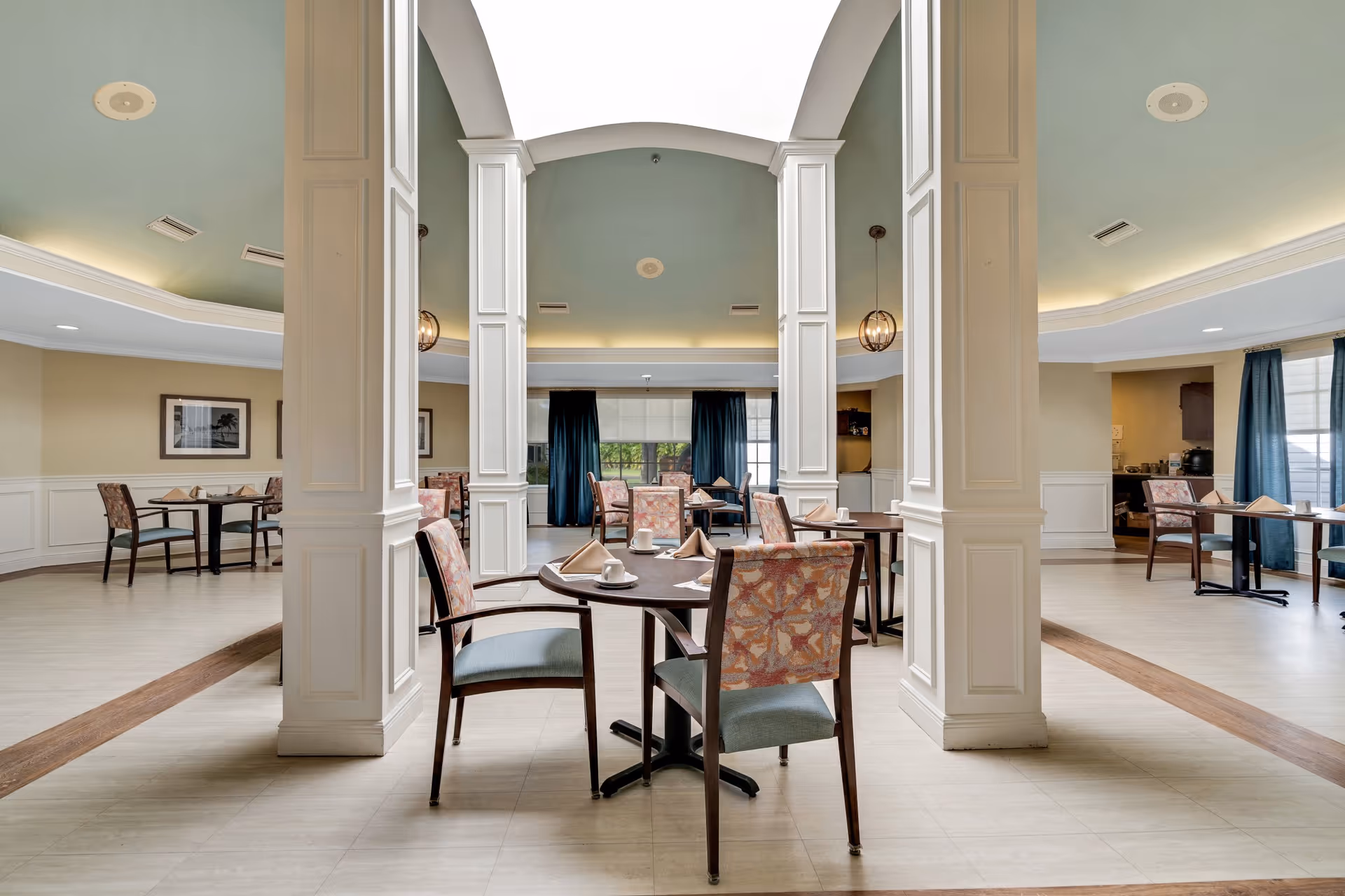 A spacious dining room with multiple round tables and chairs arranged neatly. Each table is set with napkins and cups. The room features tall white columns, a light green ceiling with recessed lighting, and large windows with dark curtains allowing natural light to enter.