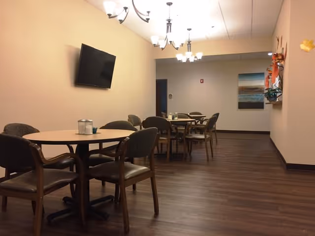 A dining area with several round tables and chairs arranged on a wooden floor. The walls are painted beige, and there are multiple ceiling light fixtures. A flat-screen TV is mounted on the left wall, and a piece of artwork is hung on the far wall. Some decorative items are visible on the right wall.