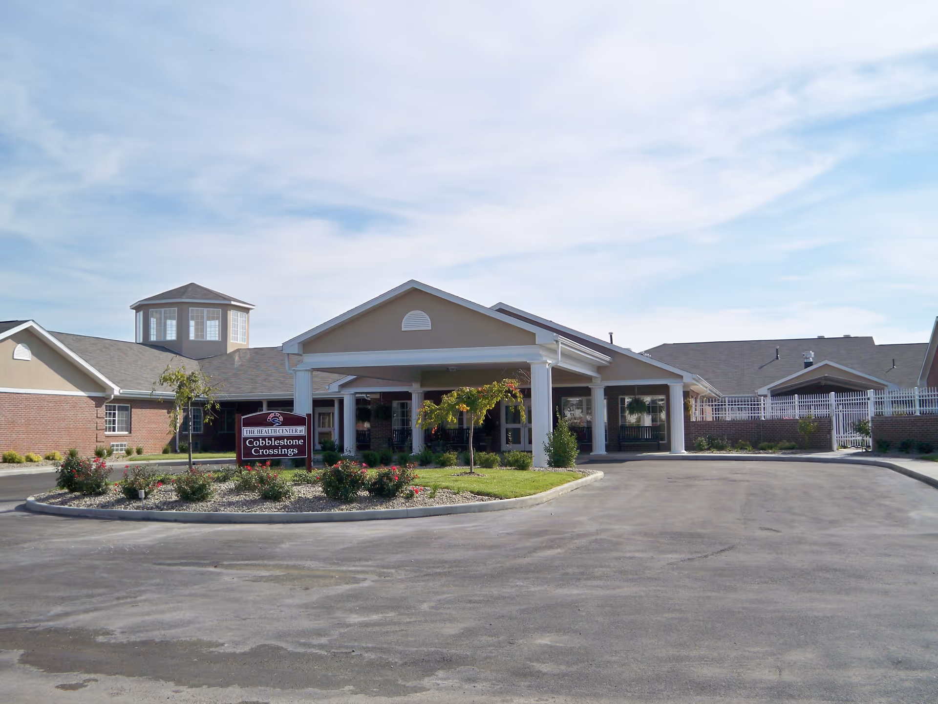 Exterior front view of Cobblestone Crossings Health Campus building with a covered entrance, landscaped garden area with small trees and bushes, and a clear sky above.
