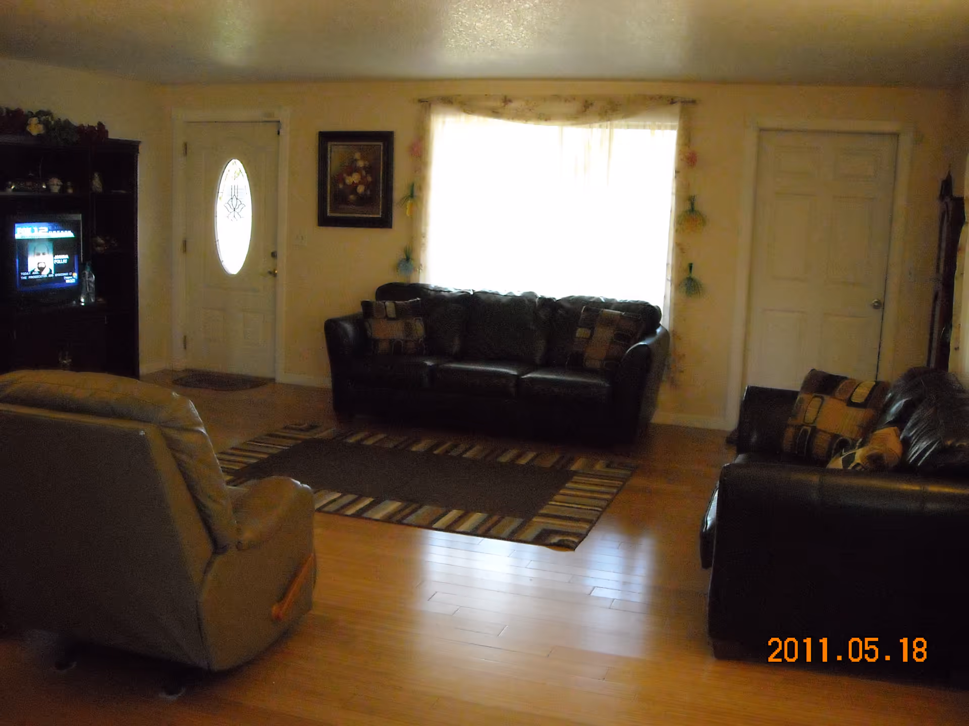 A living room with wooden flooring featuring two black leather sofas with patterned cushions, a beige recliner chair, a striped area rug, a window with sheer curtains, a white front door with an oval glass panel, a framed floral painting on the wall, and a dark wooden entertainment unit with a television displaying a news channel.