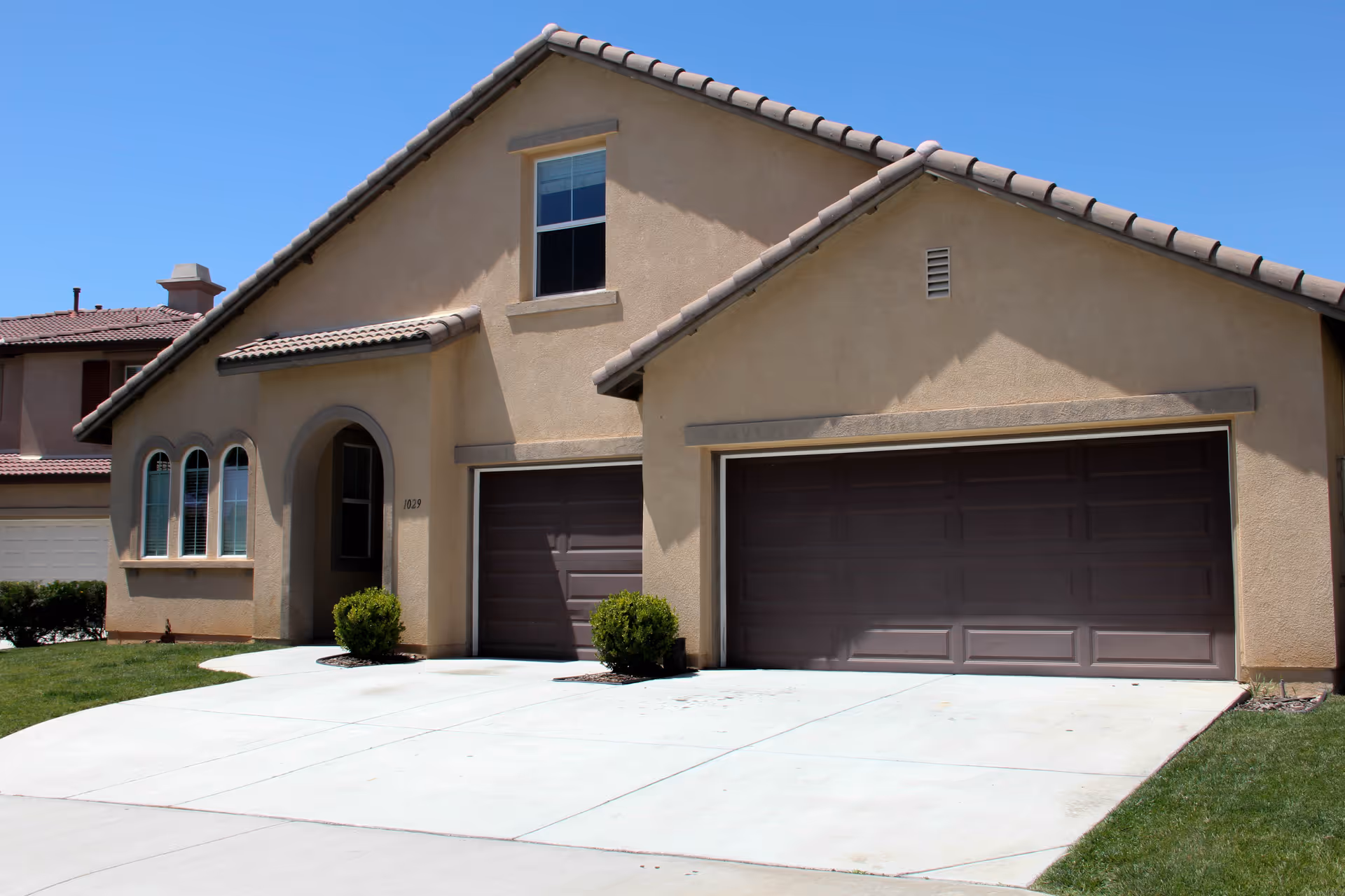 Front exterior of a beige stucco suburban house with a three-car garage and driveway under a clear blue sky.