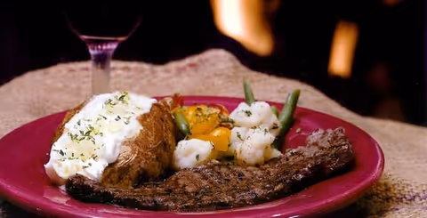 A plate with a baked potato topped with sour cream and herbs, steamed mixed vegetables including cauliflower, green beans, and carrots, and a grilled steak. A glass of red wine is partially visible in the background.