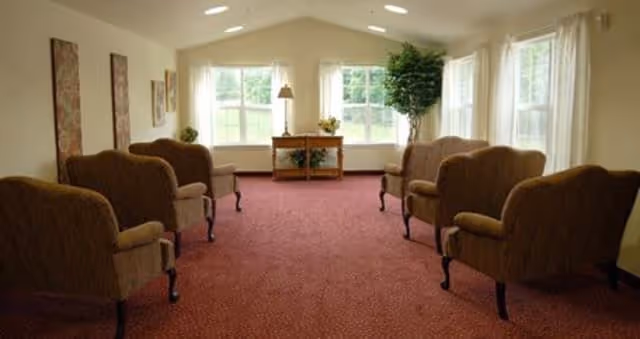 A quiet sitting area with six upholstered armchairs arranged in two rows facing each other on a red carpet. The room has large windows with white curtains letting in natural light, a small wooden table with a lamp and decorative items, and a tall green plant in the corner.