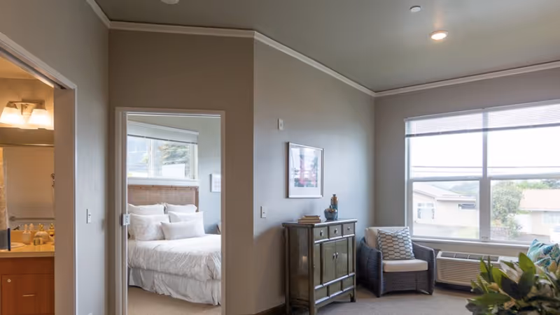 Interior view of a senior living facility room showing a cozy sitting area with a cushioned chair and a small cabinet under a framed picture. Through an open doorway, a bedroom with a neatly made bed with white linens and multiple pillows is visible. A bathroom with a lit vanity mirror is partially visible to the left. Large windows allow natural light to brighten the space.