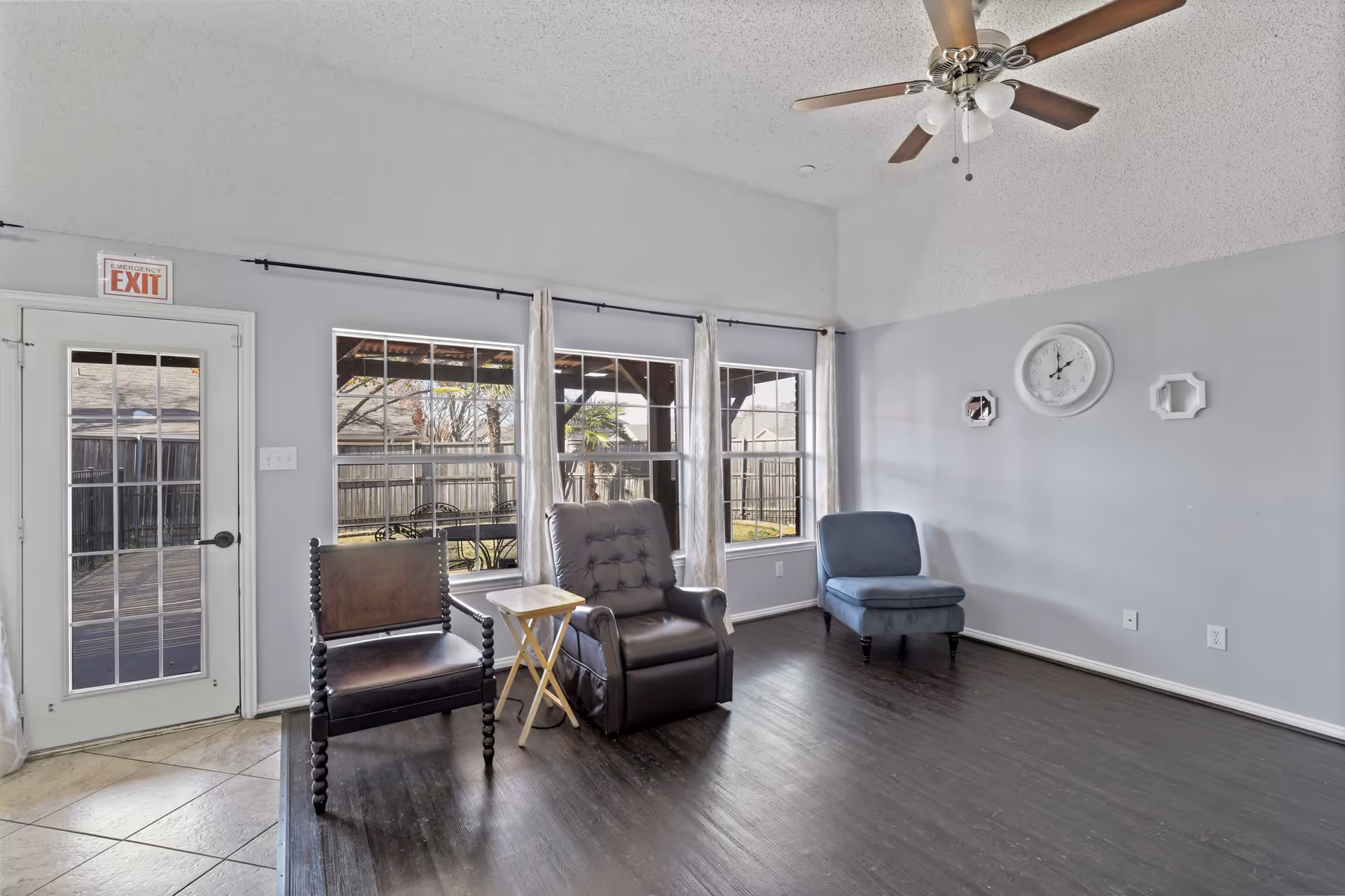 A bright living room with light gray walls and dark wood flooring. The room features a ceiling fan with lights, three chairs including a leather armchair, a wooden chair, and a blue upholstered chair. There is a small wooden side table between two of the chairs. Large windows with white curtains let in natural light and show a fenced backyard with a covered patio. An emergency exit door is visible on the left side of the room. A white clock and two decorative wall pieces hang on the right wall.