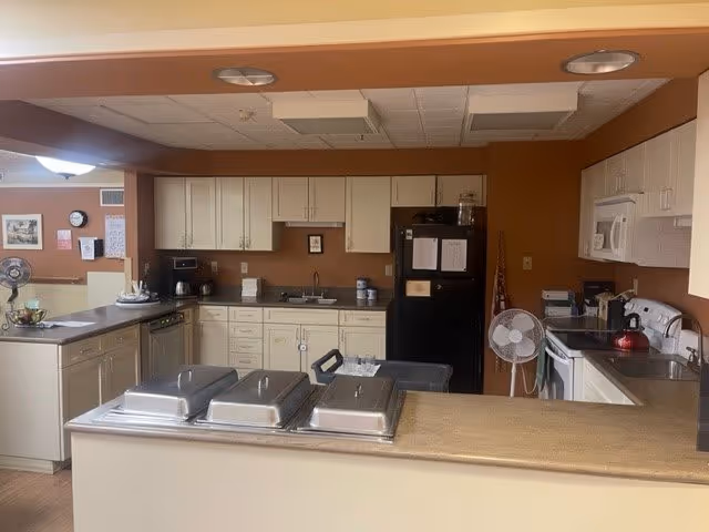 A kitchen area in a senior living facility with beige cabinets, a black refrigerator, a white microwave, a stove, and a countertop with three covered serving trays. There is a fan on the floor next to the refrigerator and various kitchen items on the counters. The walls are painted a warm brown color.