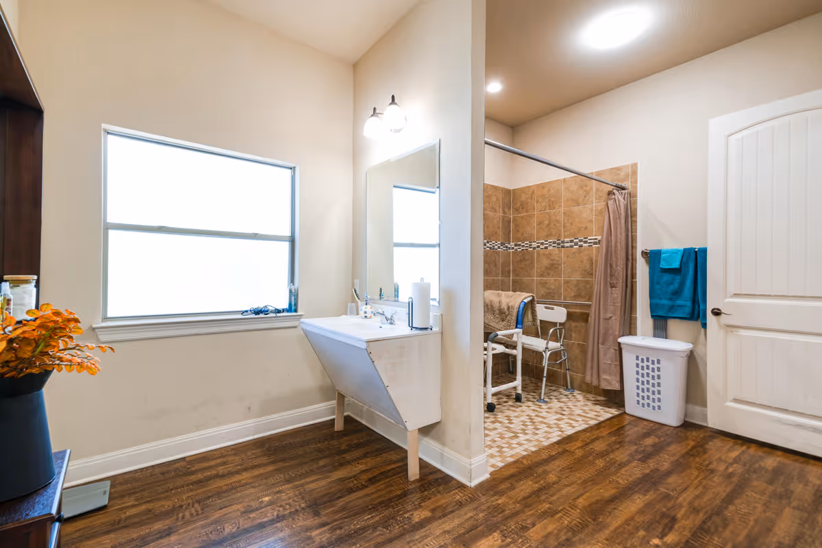 Spacious assisted-living bathroom with a sink under a mirror, a large frosted window, a tiled walk-in shower with a shower chair, and wood-look flooring.