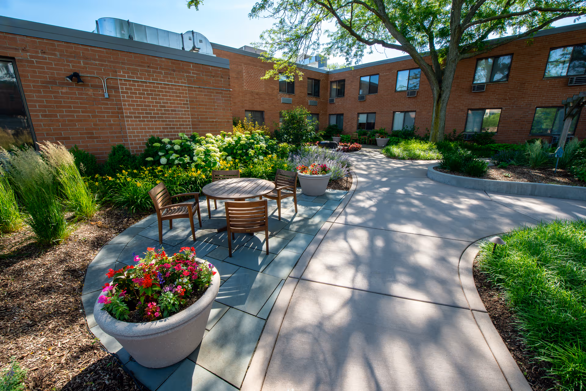 Outdoor courtyard area at Central Baptist Village featuring a round wooden table with four chairs on a stone patio surrounded by flower pots with colorful flowers, green shrubs, and trees. The courtyard is adjacent to a two-story brick building with multiple windows.