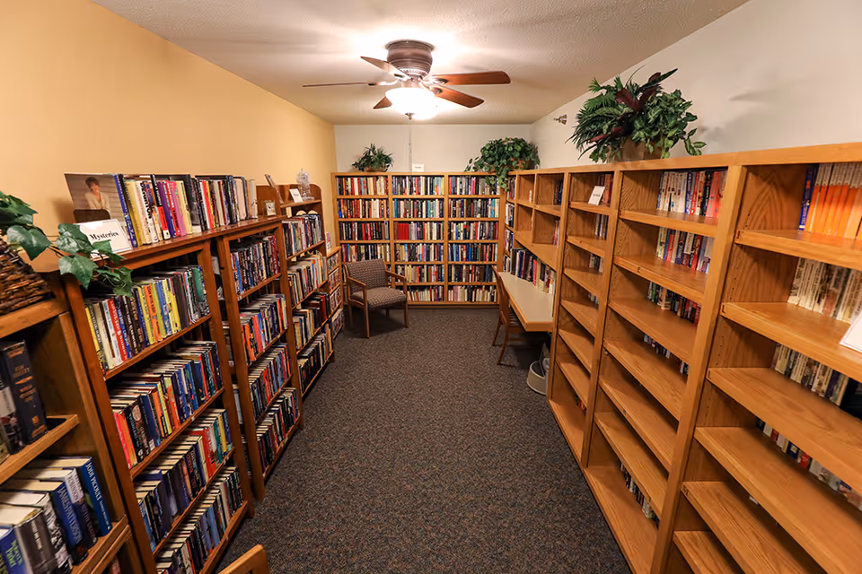 Small interior library with wooden bookshelves lining both sides, a chair and table at the far end, and a ceiling fan overhead.